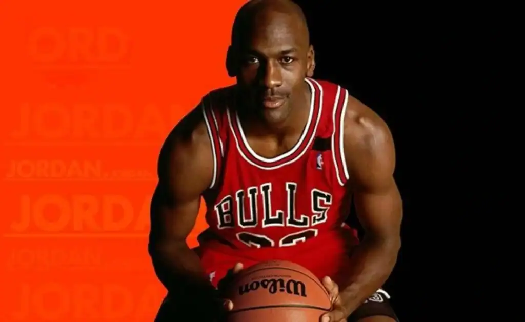 빨간 시카고 불스 유니폼을 입은 마이클 조던이 농구공을 들고 정면을 응시하는 모습, NBA 역사상 가장 위대한 선수로 평가받는 인물